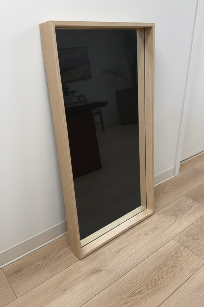 Person standing in front of a large floor mirror with wooden frame on a light wood floor.