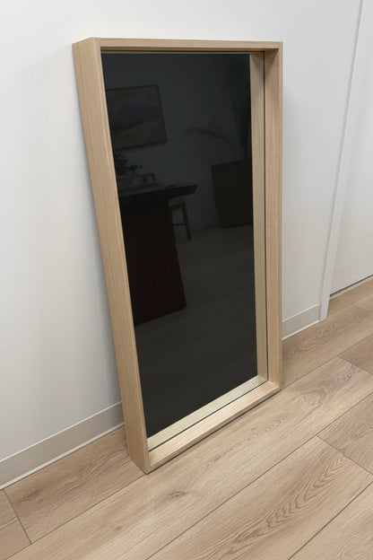 Person standing in front of a large floor mirror with wooden frame on a light wood floor.
