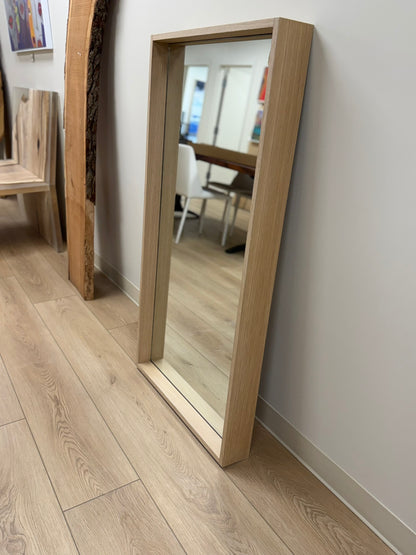Wooden floor mirror leaning against a white wall on a wooden floor.